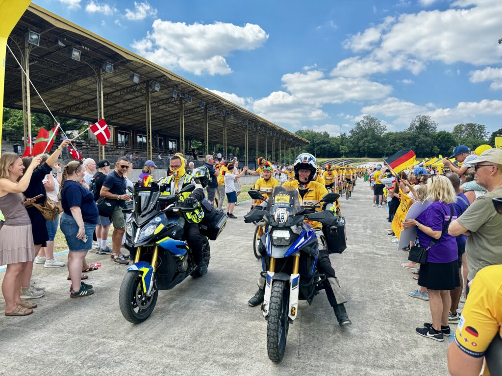 Begrüßung von Radfahrenden bei einer Großveranstaltung im Stadion: Zwei Motorradfahrer in gelber Eventkleidung fahren voran, dicht gefolgt von Radfahrerinnen und Radfahrern in gelben Trikots, während zahlreiche Zuschauer mit Deutschland- und Dänemark-Fahnen am Rand jubeln – sportliches Event mit internationaler Beteiligung und festlicher Stimmung.