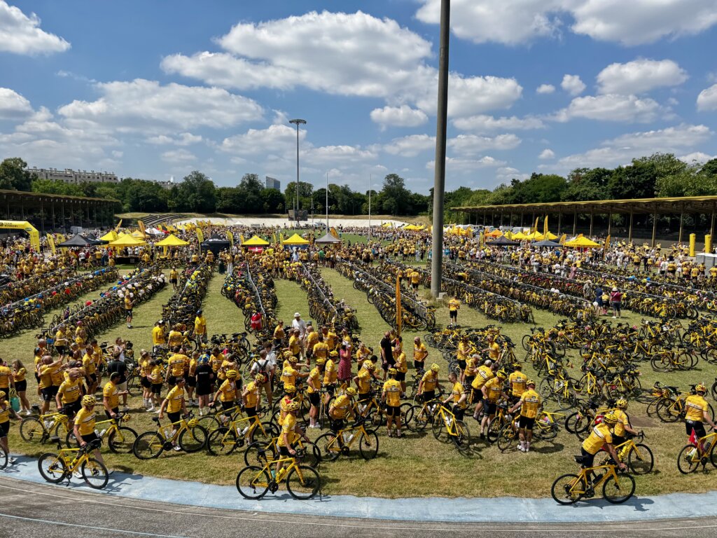 Großes Fahrradevent in einem offenen Radstadion bei sonnigem Wetter, hunderte Teilnehmende in gelben Trikots und Helmen mit gelb-schwarzen Rennrädern – dichte Reihen von Fahrrädern, zahlreiche Infostände und Pavillons im Hintergrund, sportliches Gemeinschaftserlebnis unter freiem Himmel.