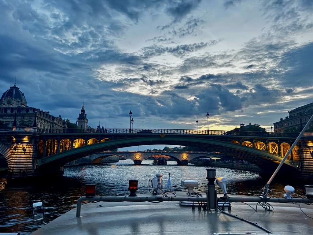 Abendliche Bootsfahrt auf der Seine in Paris mit Blick auf die beleuchtete Pont au Change und historische Gebäude bei dramatischer Wolkenstimmung im Sonnenuntergang – romantisches Stadtpanorama mit Brückenarchitektur, Wasser und französischer Hauptstadt-Atmosphäre.