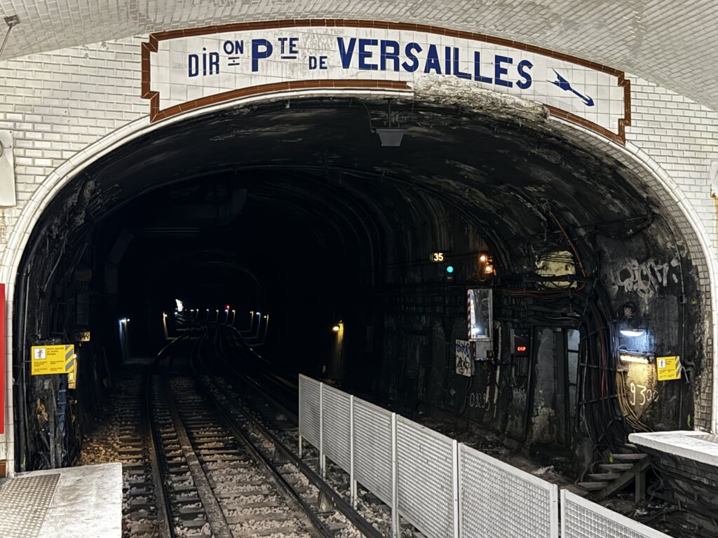 Einfahrt in einen dunklen Métro-Tunnel der Pariser U-Bahn mit dem Richtungsschild „Direction Porte de Versailles“ über dem Tunnelbogen – beleuchtet von Signalleuchten und umgeben von weißen Wandfliesen und sichtbaren Kabeln, typische Szene eines historischen Pariser Metro-Bahnhofs.