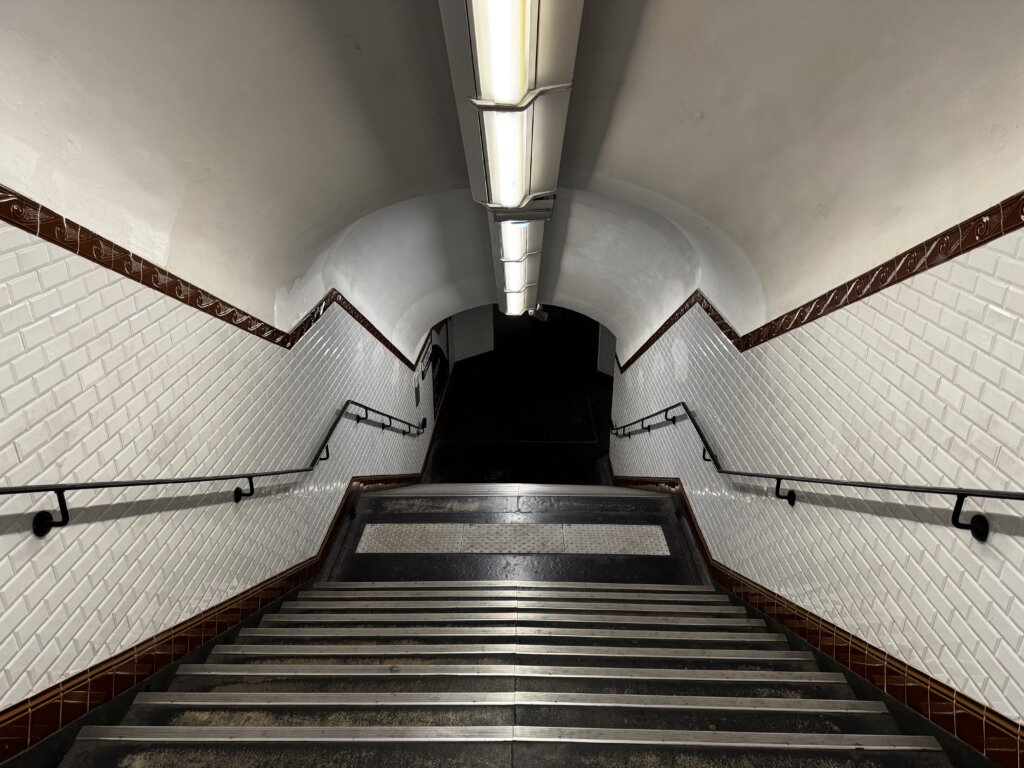 Treppenabgang in einer Pariser Metrostation mit weiß gefliesten Wänden, brauner Zierleiste und schwarzem Handlauf – beleuchtet von Neonröhren und führend in einen dunklen U-Bahn-Tunnel. Typische Architektur der Pariser Métro mit historischer Fliesenoptik und urbanem Flair.