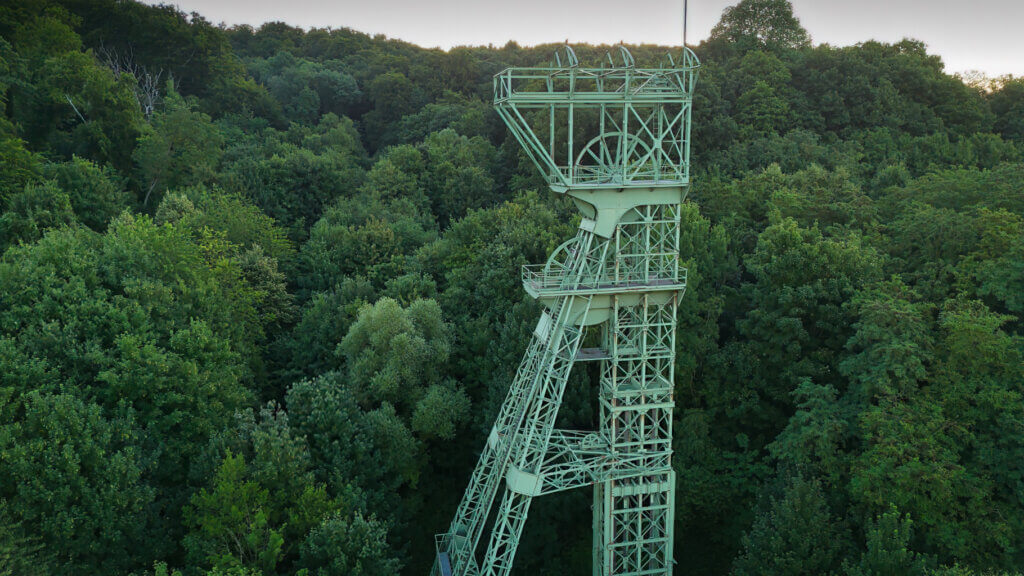 Detailaufnahme des Förderturms der ehemaligen Zeche Carl Funke in Essen, eingerahmt von dichtem, grünem Laubwald. Die grüne Stahlkonstruktion mit markantem Seilscheiben-Gerüst ragt filigran über die Baumwipfel hinaus und erinnert an die industrielle Vergangenheit des Ruhrgebiets inmitten der Naturkulisse am Baldeneysee.