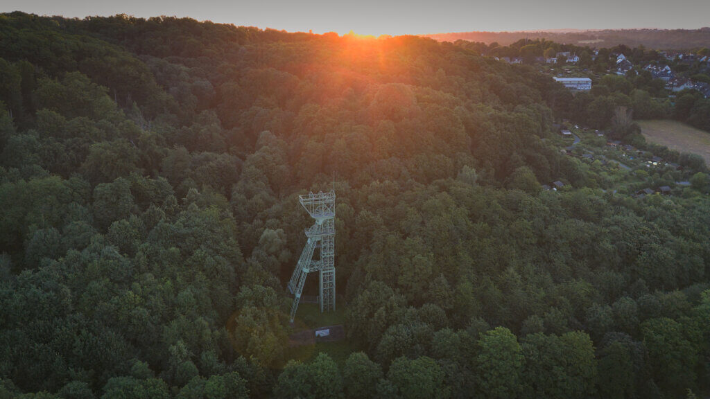 Luftaufnahme des Förderturms der Zeche Carl Funke in Essen bei Sonnenuntergang. Die grüne Stahlkonstruktion ragt aus einem dichten Waldgebiet, während die Sonne hinter dem Hügel am Horizont versinkt und warmes Licht über das Ruhrtal legt. Rechts im Bild erkennt man Kleingartenanlagen und Wohnhäuser am Waldrand.