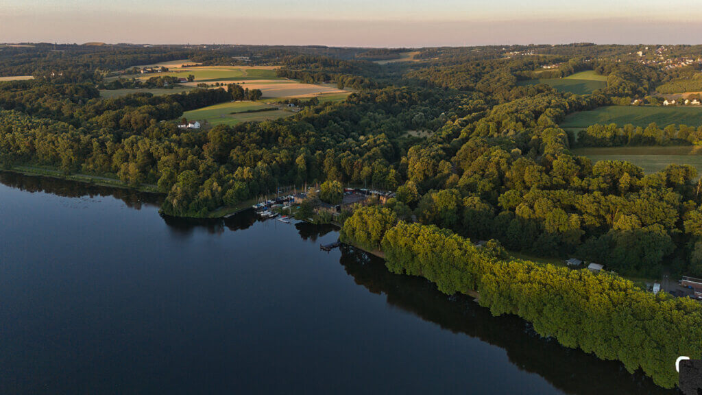 Luftaufnahme des Baldeneysees in Essen mit Blick auf das Schloss Baldeney, eingebettet in einen dichten Wald am Ufer. Rund um die historische Anlage liegen Segelboote an einem kleinen Yachthafen. Im Hintergrund erstreckt sich eine sanfte Hügellandschaft mit Feldern, Wiesen und einzelnen Höfen – typisch für die ländliche Umgebung des südlichen Ruhrgebiets bei Sonnenaufgang.