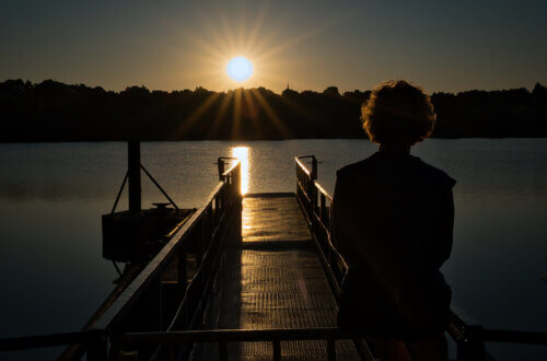 Silhouette einer Person bei Sonnenaufgang auf einem Steg am Baldeneysee in Essen. Die untergehende Sonne wirft goldenes Licht auf das Wasser und den Metallsteg, während sich die Umrisse des gegenüberliegenden Ufers abzeichnen. Die Szene vermittelt Ruhe, Reflexion und Naturverbundenheit inmitten des Ruhrgebiets. Baldeneysee Essen Sonnenaufgang