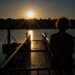 Silhouette einer Person bei Sonnenaufgang auf einem Steg am Baldeneysee in Essen. Die untergehende Sonne wirft goldenes Licht auf das Wasser und den Metallsteg, während sich die Umrisse des gegenüberliegenden Ufers abzeichnen. Die Szene vermittelt Ruhe, Reflexion und Naturverbundenheit inmitten des Ruhrgebiets. Baldeneysee Essen Sonnenaufgang