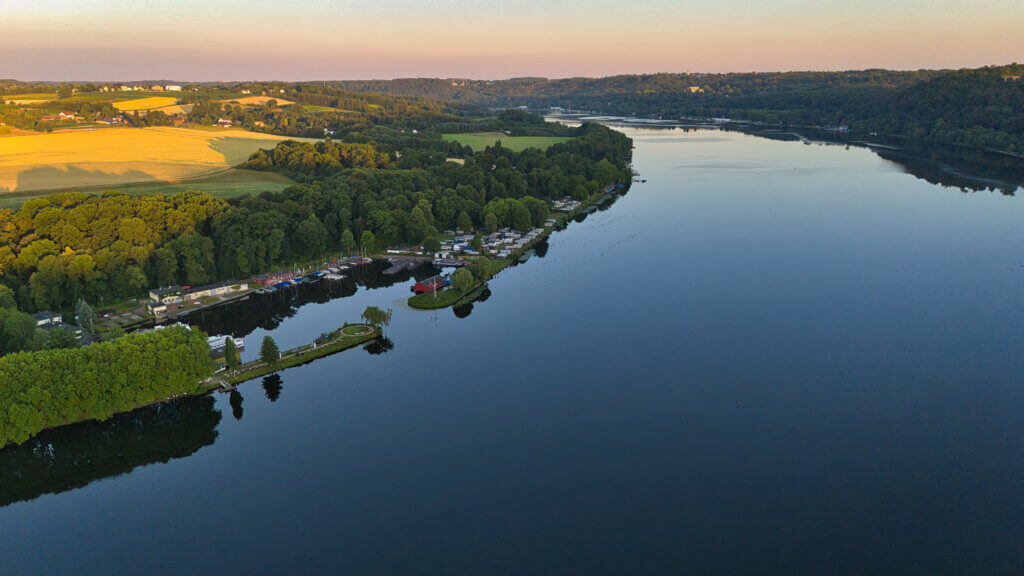 Panoramaaufnahme des Baldeneysees in Essen bei Sonnenaufgang. Der spiegelglatte See erstreckt sich entlang bewaldeter Ufer und idyllischer Bootsanlagen. Links im Bild ein kleiner Hafen mit Segelbooten, Clubhäusern und einem Rundweg auf einer schmalen Landzunge. Im Hintergrund liegen goldene Felder, Wälder und sanfte Hügel der Essener Stadtgrenze.