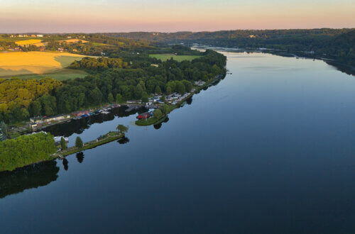 Panoramaaufnahme des Baldeneysees in Essen bei Sonnenaufgang. Der spiegelglatte See erstreckt sich entlang bewaldeter Ufer und idyllischer Bootsanlagen. Links im Bild ein kleiner Hafen mit Segelbooten, Clubhäusern und einem Rundweg auf einer schmalen Landzunge. Im Hintergrund liegen goldene Felder, Wälder und sanfte Hügel der Essener Stadtgrenze.