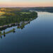 Panoramaaufnahme des Baldeneysees in Essen bei Sonnenaufgang. Der spiegelglatte See erstreckt sich entlang bewaldeter Ufer und idyllischer Bootsanlagen. Links im Bild ein kleiner Hafen mit Segelbooten, Clubhäusern und einem Rundweg auf einer schmalen Landzunge. Im Hintergrund liegen goldene Felder, Wälder und sanfte Hügel der Essener Stadtgrenze.