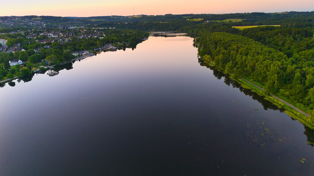 Weitwinkel-Luftaufnahme des Baldeneysees in Essen bei Sonnenaufgang. Das ruhige Wasser spiegelt den pastellfarbenen Himmel, während am linken Ufer Wohnhäuser, Segelboothäfen und Grünanlagen zu sehen sind. Rechts verläuft ein idyllischer Uferweg entlang dichter Wälder. Im Hintergrund erstrecken sich sanfte Hügel und Felder im südlichen Ruhrgebiet.