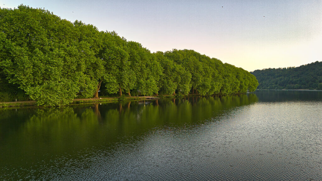 Weitwinkel-Luftaufnahme des Baldeneysees in Essen bei Sonnenaufgang. Das ruhige Wasser spiegelt den pastellfarbenen Himmel, während am linken Ufer Wohnhäuser, Segelboothäfen und Grünanlagen zu sehen sind. Rechts verläuft ein idyllischer Uferweg entlang dichter Wälder. Im Hintergrund erstrecken sich sanfte Hügel und Felder im südlichen Ruhrgebiet.