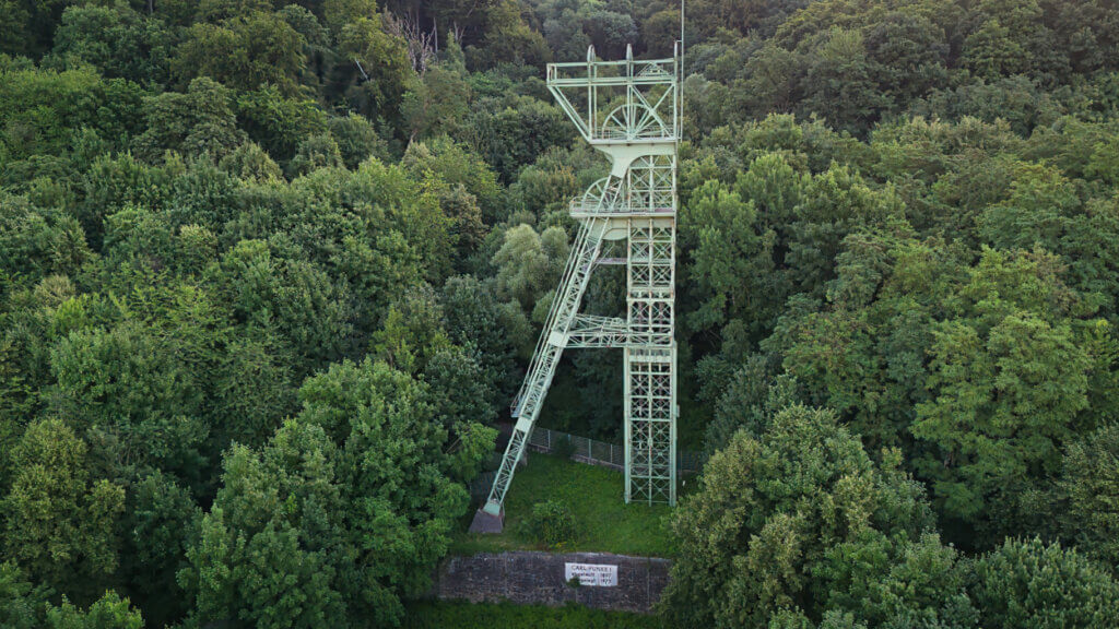 Nahaufnahme des Förderturms der ehemaligen Zeche Carl Funke in Essen, umgeben von dichtem Wald. Die grüne Stahlkonstruktion ragt filigran aus dem Blätterdach und ist gut erhalten. Am unteren Bildrand ist eine Steinmauer mit der Inschrift „Carl Funke 1, Abgeteuft 1889, Stillgelegt 1973“ sichtbar – ein Denkmal der Ruhrgebiet-Industriegeschichte mitten im Grünen.