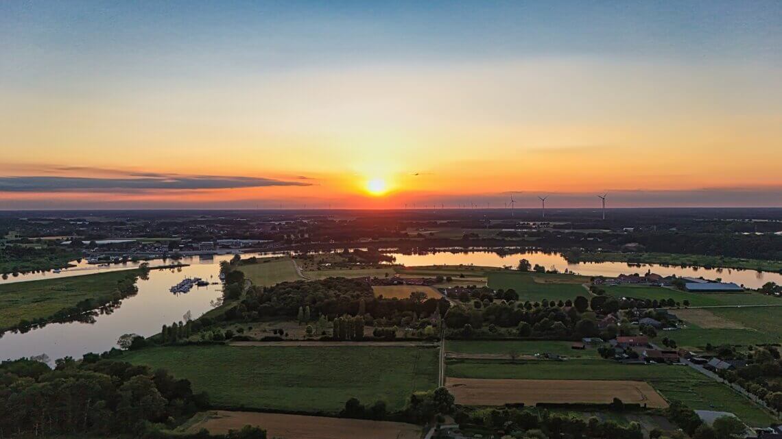 Luftaufnahme eines Sonnenuntergangs über einer ländlichen Flusslandschaft mit Wiesen, Feldern, Seen und Windrädern am Horizont – stimmungsvolle Abendstimmung im Sommer.