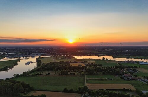 Luftaufnahme eines Sonnenuntergangs über einer ländlichen Flusslandschaft mit Wiesen, Feldern, Seen und Windrädern am Horizont – stimmungsvolle Abendstimmung im Sommer.