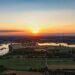 Luftaufnahme eines Sonnenuntergangs über einer ländlichen Flusslandschaft mit Wiesen, Feldern, Seen und Windrädern am Horizont – stimmungsvolle Abendstimmung im Sommer.