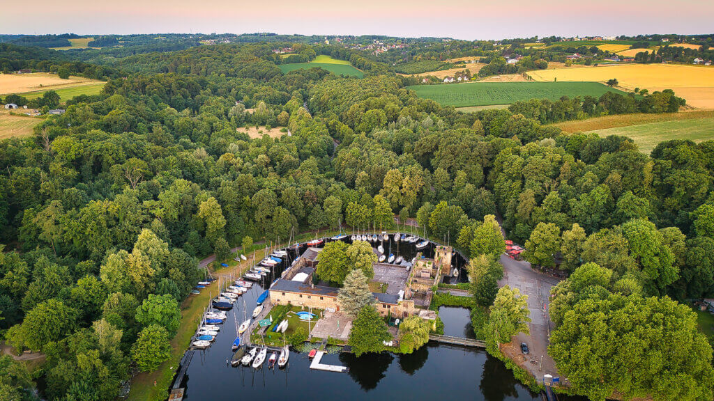 Weitwinklige Luftaufnahme des historischen Wasserschlosses Baldeney in Essen, eingebettet in dichten Mischwald am Baldeneysee. Der Yachthafen mit zahlreichen Segelbooten liegt in einem ringförmigen Hafenbecken, umgeben von alten Schlossmauern. Im Hintergrund erstrecken sich sanfte Hügel, Felder und das ländliche Ruhrgebiet bei Sonnenaufgang