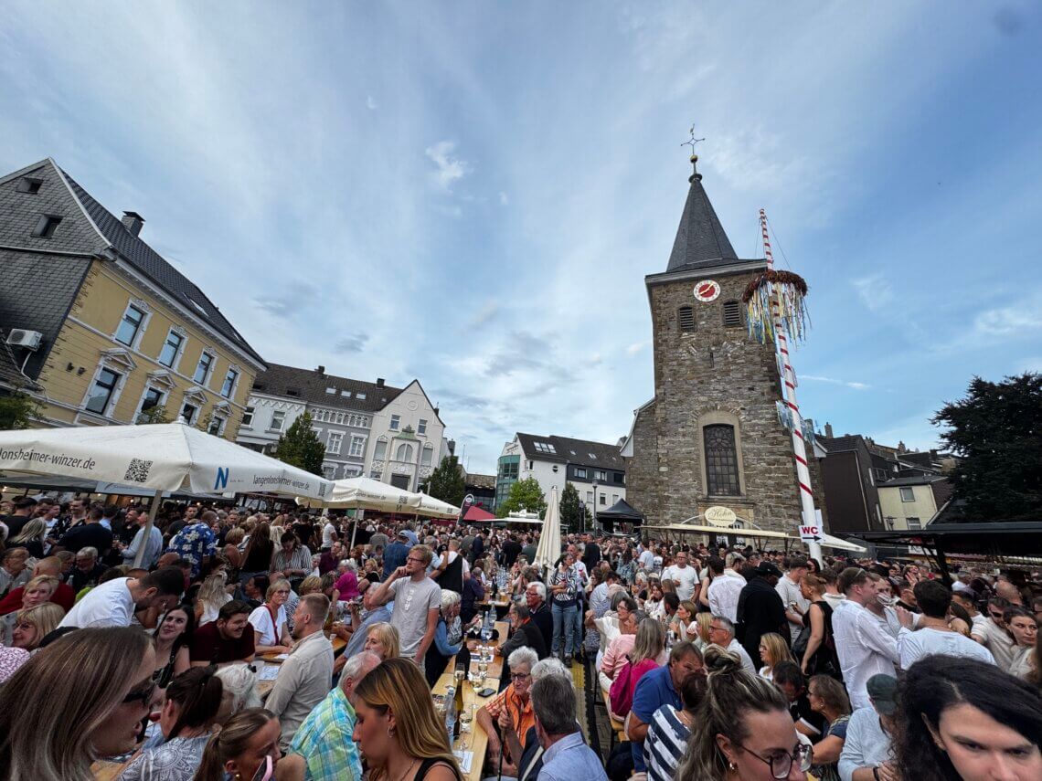 Weinfest in Velbert mit Menschenmengen vor Kirchturm bei Sonnenschein.