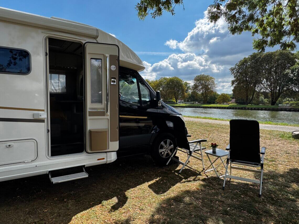 Wohnmobil am Flussufer mit geöffnetem Eingang, davor zwei Campingstühle und ein kleiner Tisch unter Bäumen, sonniger Sommertag mit blauem Himmel und Wolken