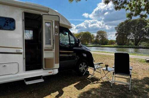 Wohnmobil am Flussufer mit geöffnetem Eingang, davor zwei Campingstühle und ein kleiner Tisch unter Bäumen, sonniger Sommertag mit blauem Himmel und Wolken
