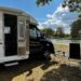 Wohnmobil am Flussufer mit geöffnetem Eingang, davor zwei Campingstühle und ein kleiner Tisch unter Bäumen, sonniger Sommertag mit blauem Himmel und Wolken