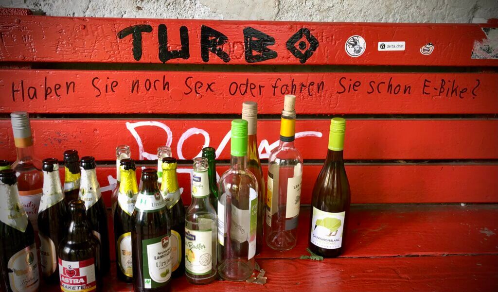 Rote Parkbank mit schwarzer Aufschrift „Haben Sie noch Sex oder fahren Sie schon E-Bike?“ und zahlreichen leeren Alkoholflaschen davor. Streettypografie