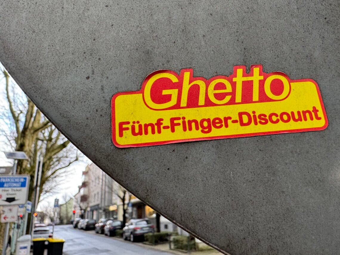 Aufkleber mit dem Schriftzug „Ghetto Fünf-Finger-Discount“ auf grauem Verkehrsschild in urbaner Straße. Fünf-Finger-Philosophie.