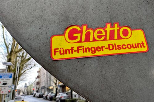 Aufkleber mit dem Schriftzug „Ghetto Fünf-Finger-Discount“ auf grauem Verkehrsschild in urbaner Straße. Fünf-Finger-Philosophie.