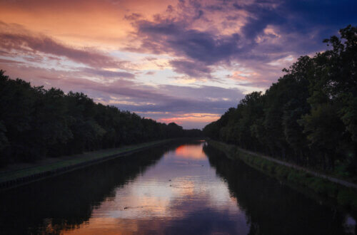 Abendstimmung. Der Mittelland-Kanal fließt zwischen zwei dicht bewaldeten Ufern. Der Himmel ist in kräftige Farben getaucht – von Orange über Rosa bis Dunkelblau. Die Wasseroberfläche reflektiert die Farben. Einige Enten schwimmen in der Ferne.
