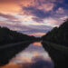 Abendstimmung. Der Mittelland-Kanal fließt zwischen zwei dicht bewaldeten Ufern. Der Himmel ist in kräftige Farben getaucht – von Orange über Rosa bis Dunkelblau. Die Wasseroberfläche reflektiert die Farben. Einige Enten schwimmen in der Ferne.