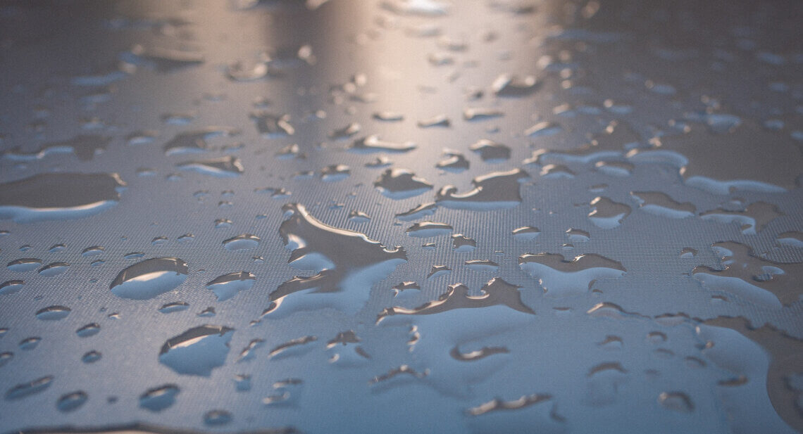 Großflächige Aufnahme von Wasserperlen auf einer glatten, leicht strukturierten Fläche mit bläulichem Schimmer – natürliche Spiegelungen bei diffusem Licht. Projekt52: Regen