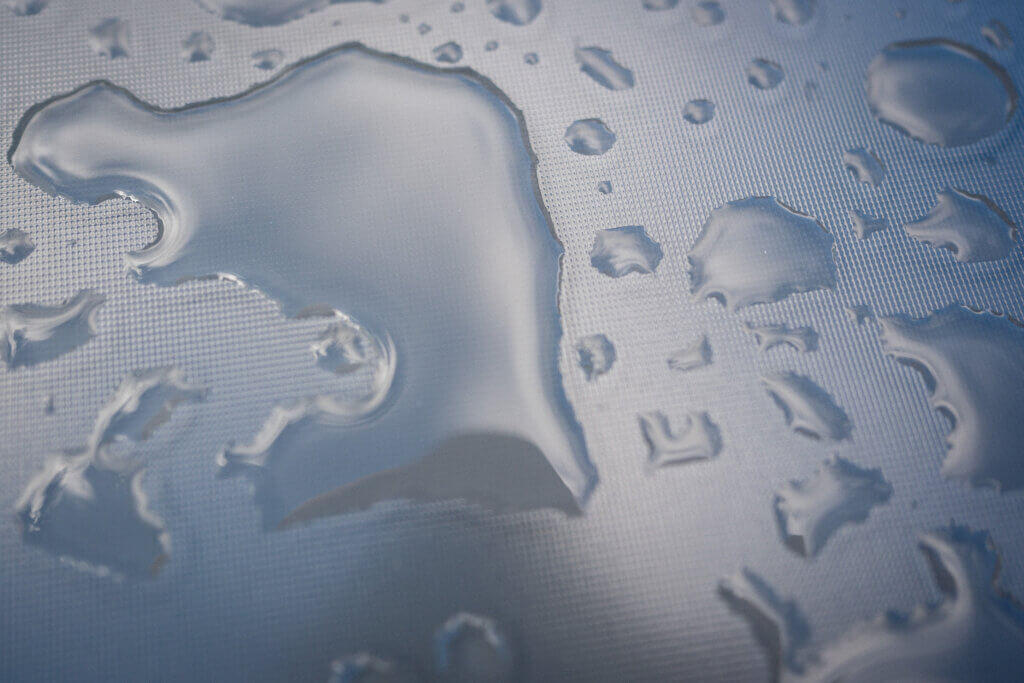 Makroaufnahme großer und kleiner Wassertropfen auf einer hellen Oberfläche mit feiner Rillenstruktur – klare Formen und weiches Licht sorgen für plastische Wirkung. Projekt52: Regen