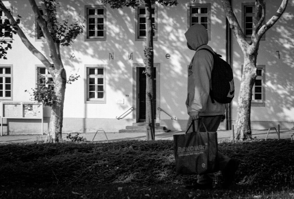 Person mit Kapuzenpullover und Einkaufstasche läuft an einem Amtsgebäude vorbei, Schatten von Bäumen fallen auf die Fassade. Streetfotografie. Streetfotografie im Ruhestand
