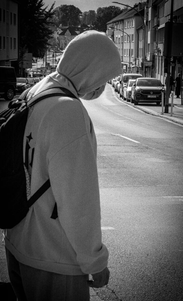 Mensch mit Kapuze und Rucksack steht an einer Straße in der Stadt und blickt in Richtung des Verkehrs. Streetfotografie. Streetfotografie im Ruhestand.