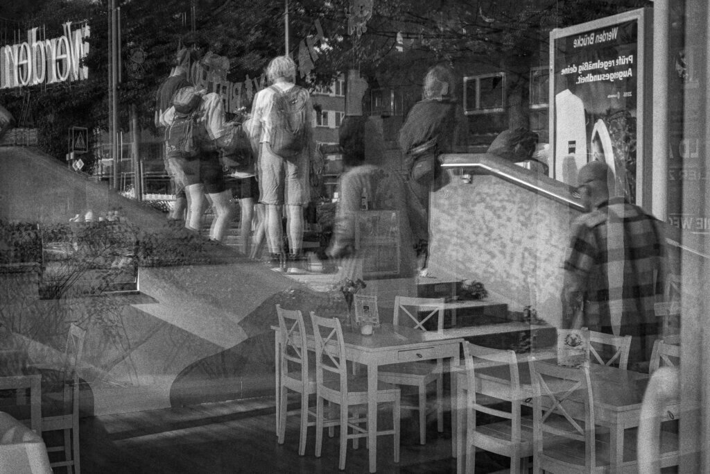 Straßenszene spiegelt sich in einer Fensterscheibe eines Cafés mit leeren Stühlen, Menschen stehen draußen in einer Gruppe. Streetfotografie im Ruhestand