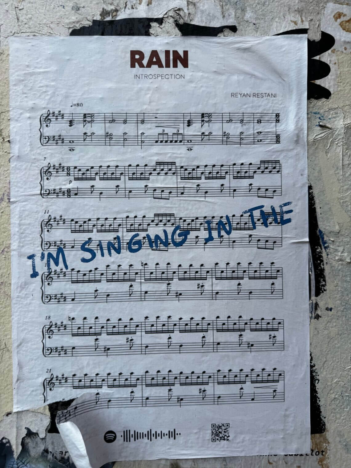 Ein an einer verwitterten Wand angebrachtes Plakat zeigt ein Notenblatt mit dem Titel „RAIN – Introspection“ von Reyan Restani. In großen blauen Buchstaben ist quer über die Noten handschriftlich der Satz „I’M SINGING IN THE“ geschrieben. Unten auf dem Plakat befindet sich ein Spotify-Code und ein QR-Code.