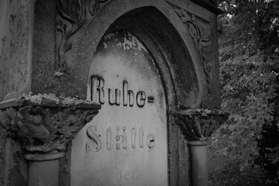 Schwarz-weiß-Aufnahme eines stark verwitterten Grabsteins mit der Inschrift „Ruhe-Stätte“. Der Stein ist in ein halbrundes, gotisch inspiriertes Steinportal eingelassen, flankiert von dekorativen Säulen mit Eichenlaub-Kapitellen. Die Oberfläche ist von Moos und Flechten überzogen, Putz blättert ab. Im Hintergrund unscharf: dichtes Laub eines alten Baumes. Die Szene wirkt melancholisch und von der Zeit gezeichnet. Stiller Park Velbert