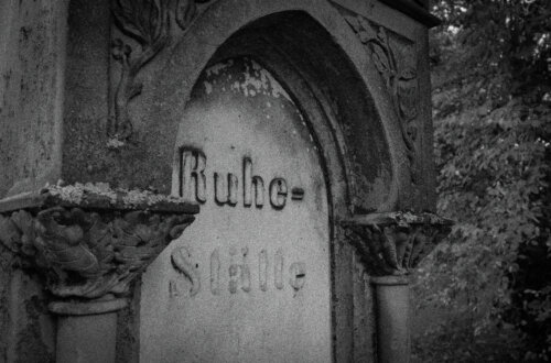 Schwarz-weiß-Aufnahme eines stark verwitterten Grabsteins mit der Inschrift „Ruhe-Stätte“. Der Stein ist in ein halbrundes, gotisch inspiriertes Steinportal eingelassen, flankiert von dekorativen Säulen mit Eichenlaub-Kapitellen. Die Oberfläche ist von Moos und Flechten überzogen, Putz blättert ab. Im Hintergrund unscharf: dichtes Laub eines alten Baumes. Die Szene wirkt melancholisch und von der Zeit gezeichnet. Stiller Park Velbert