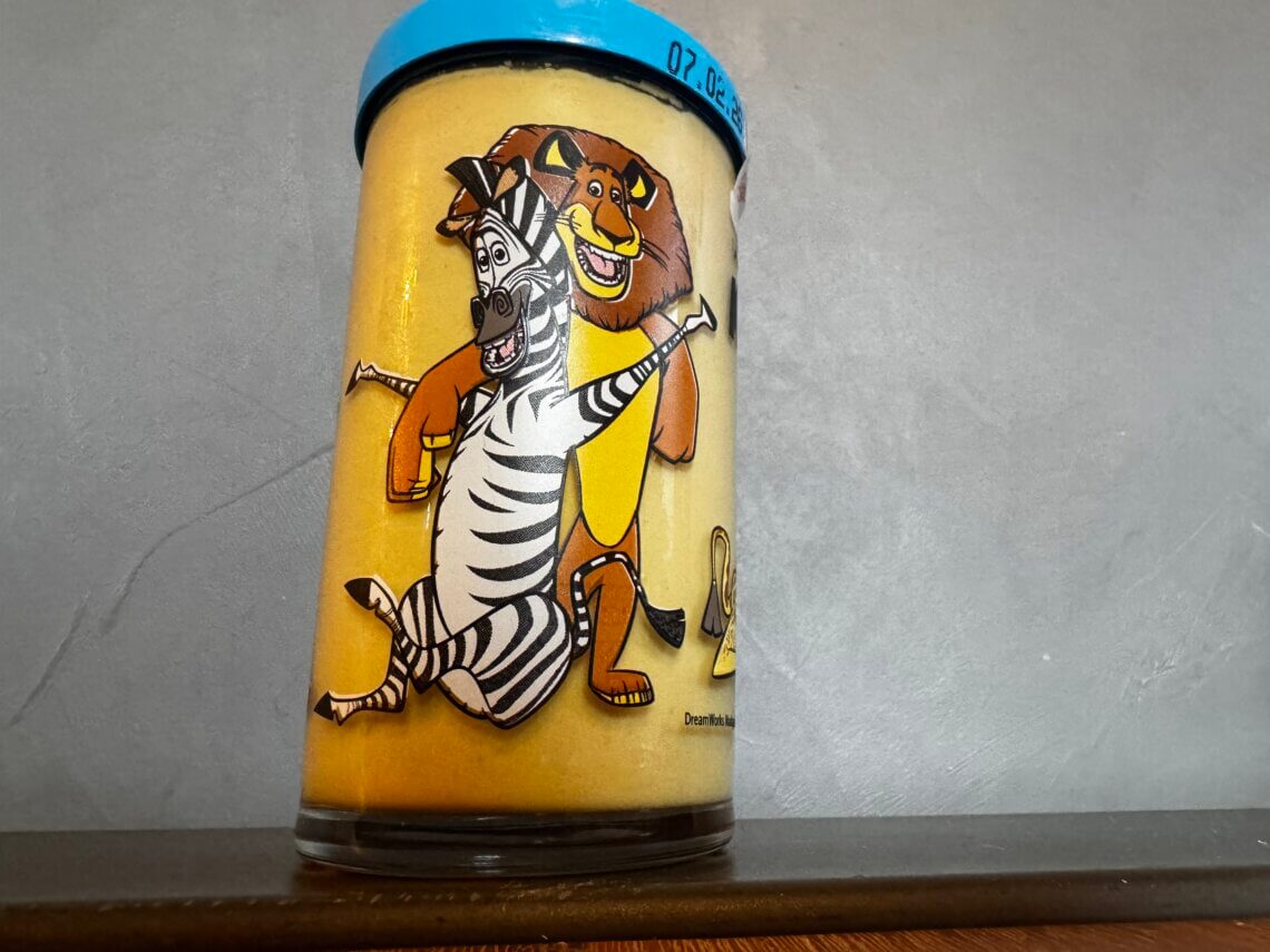 Kindheitserinnerung in Glas. Volles Senfglas mit Madagascar-Figuren Zebra Marty und Löwe Alex vor grauer Wand
