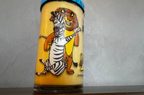 Kindheitserinnerung in Glas. Volles Senfglas mit Madagascar-Figuren Zebra Marty und Löwe Alex vor grauer Wand