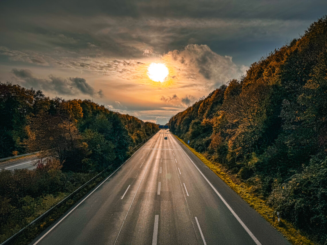 Leere Autobahn im Licht der untergehenden Sonne, umgeben von bunt gefärbtem Herbstwald – stimmungsvolles Foto einer herbstlichen Reise in den Ruhestand.