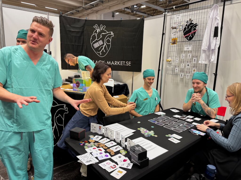 Messestand des Spiels „Black Market“ mit Spielleiter*innen in OP-Kleidung – schwarzer Humor trifft Kartenspiel auf der SPIEL Essen.