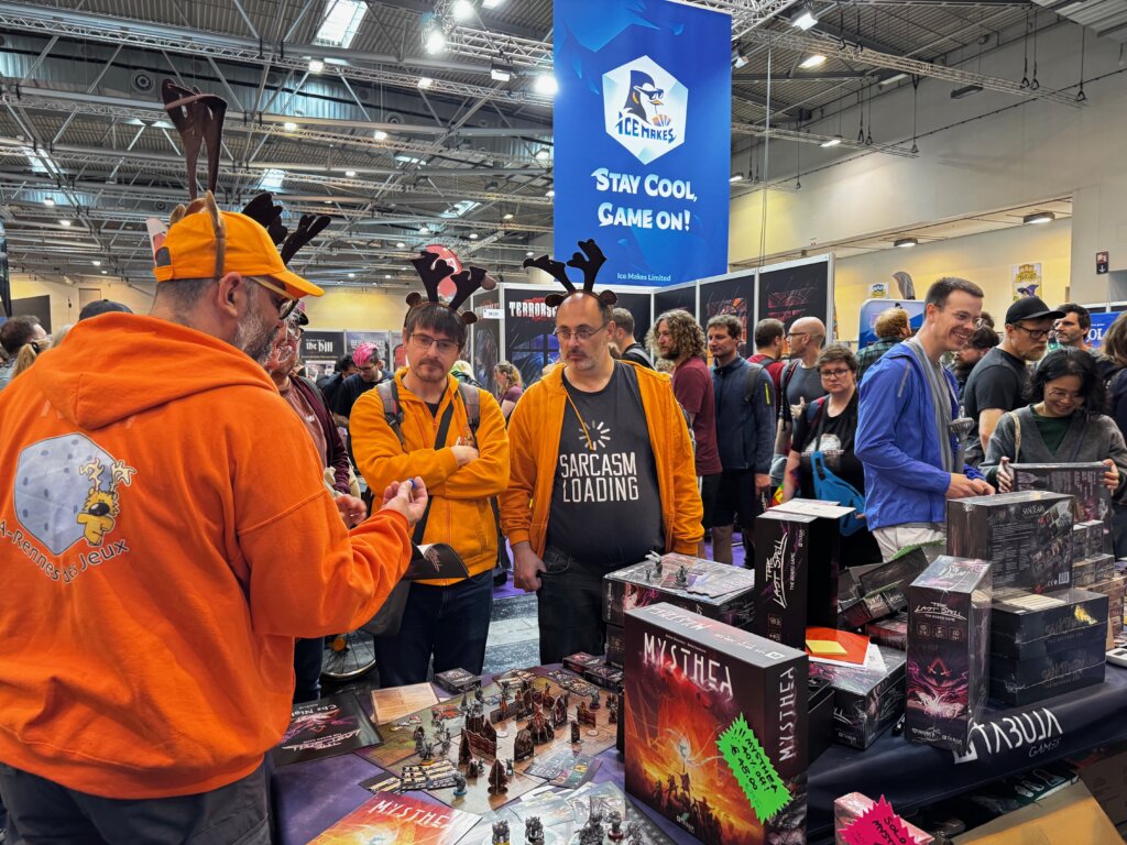 Gut gelaunte Männer in orangefarbenen Outfits mit Rentiergeweihen am Stand von Tabula Games auf der SPIEL Essen Messe.