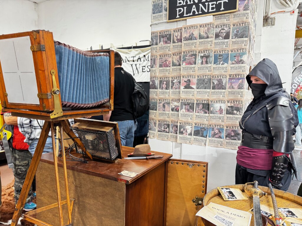 Nostalgische Fotokulisse im Western-Stil – Cosplayerin mit Kapuze vor „Wanted“-Wand und antiker Kamera auf der SPIEL Essen.
