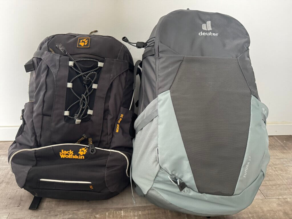 Zwei Rucksäcke nebeneinander: links ein schwarzer Jack-Wolfskin-Rucksack mit 22 Litern, rechts ein grauer Deuter-Rucksack mit 27 Litern Volumen. Beide stehen auf einem Holzfußboden vor weißer Wand.Rom Reise mit Handgepäck