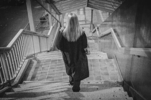 Frau mit langen blonden Haaren steigt Betontreppe hinauf, Schwarzweiß-Streetfotografie, Rückenansicht, urbane Architektur