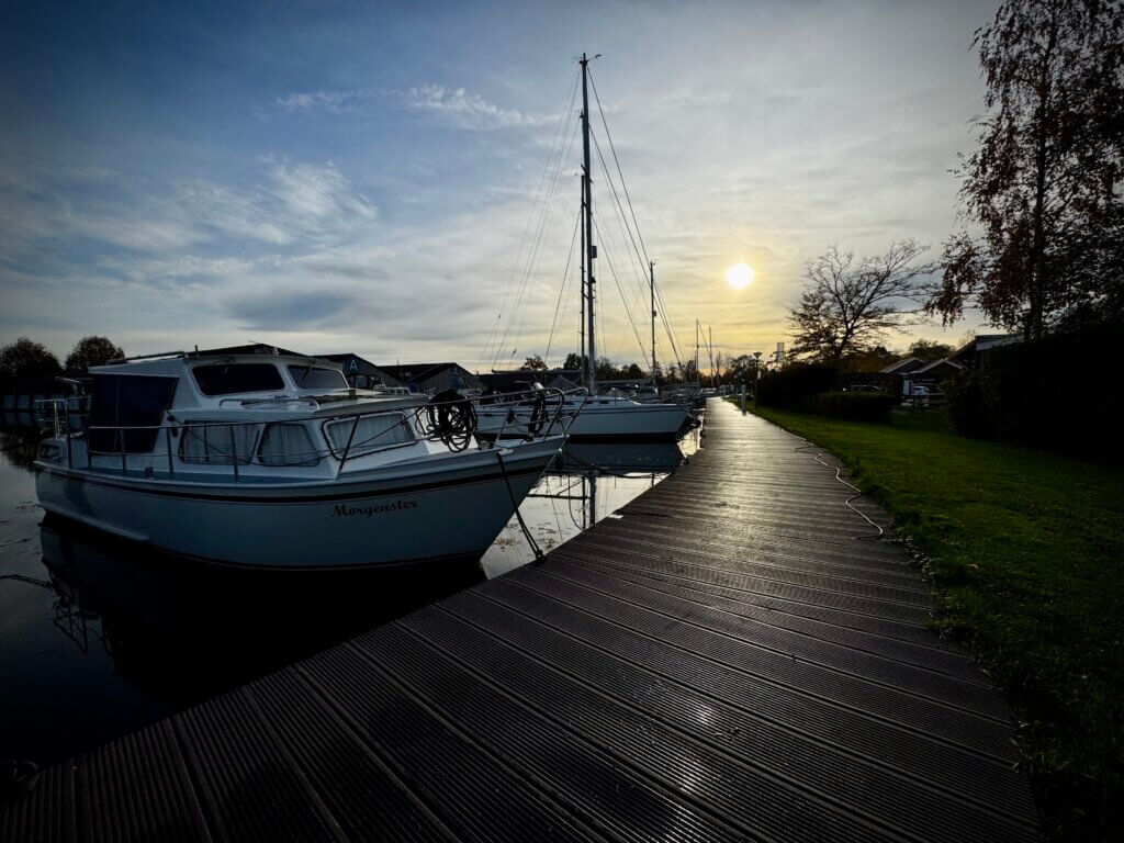 Weiße Boote liegen still im Wasser an einem Holzsteg in Sneek, die tief stehende Sonne spiegelt sich im Wasser. Rechts führt ein Weg am Ufer entlang, herbstliche Bäume rahmen das Bild. Friedlicher Moment am Wasser. Wohnmobilreise Sneek Niederlande.
