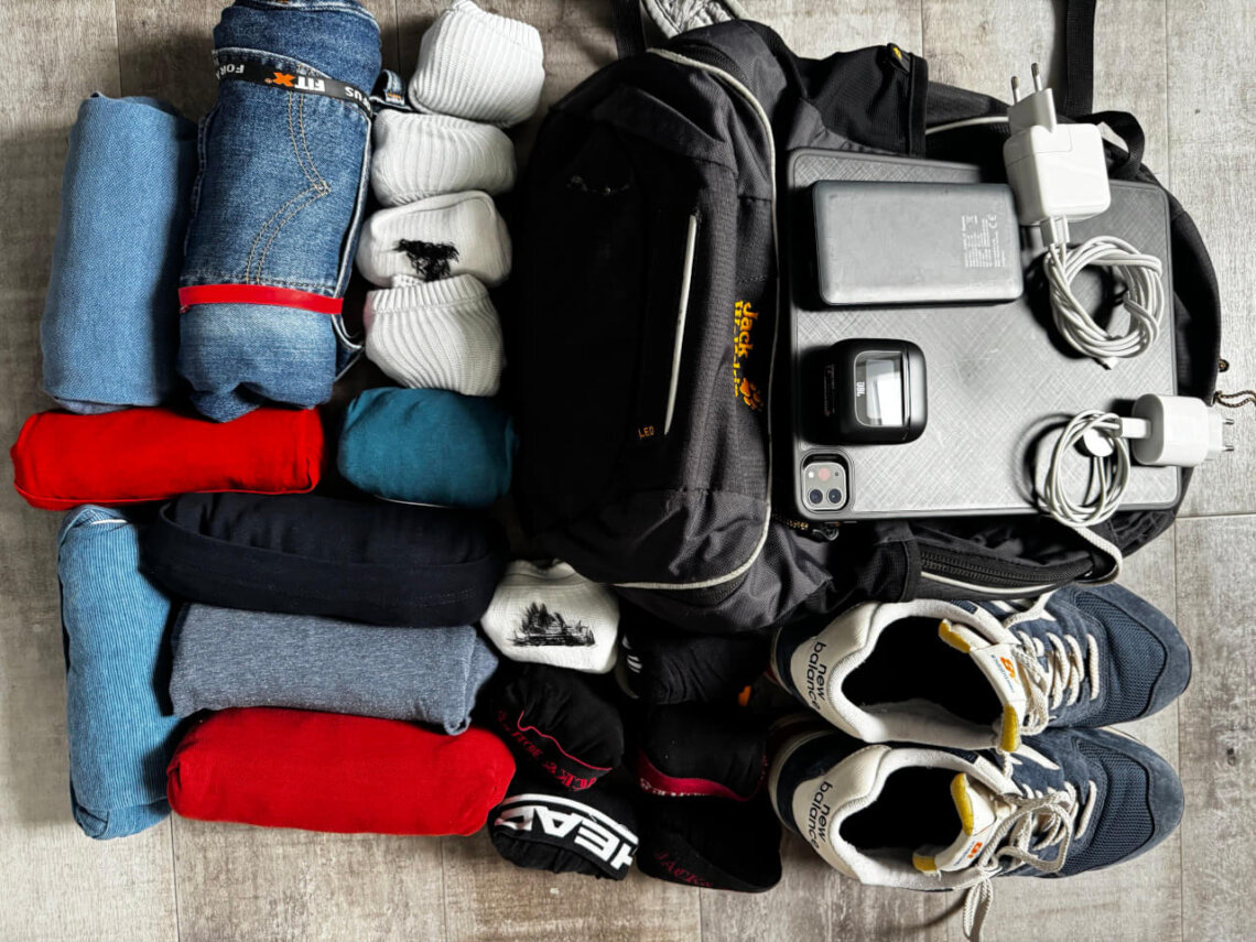Ein 22 l Reiserucksack mit daneben ausgebreiteter Kleidung und Technik: zusammengerollte T-Shirts, Hosen, Socken, ein Paar Sneaker, Ladegeräte, Tablet, Powerbank und ein kleiner Kopfhörer liegen ordentlich sortiert auf einem Boden. Rom Reise mit Handgepäck.