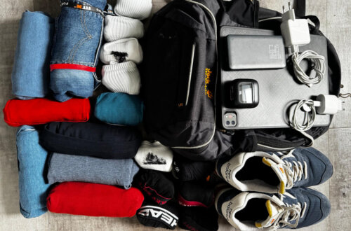 Ein 22 l Reiserucksack mit daneben ausgebreiteter Kleidung und Technik: zusammengerollte T-Shirts, Hosen, Socken, ein Paar Sneaker, Ladegeräte, Tablet, Powerbank und ein kleiner Kopfhörer liegen ordentlich sortiert auf einem Boden. Rom Reise mit Handgepäck.