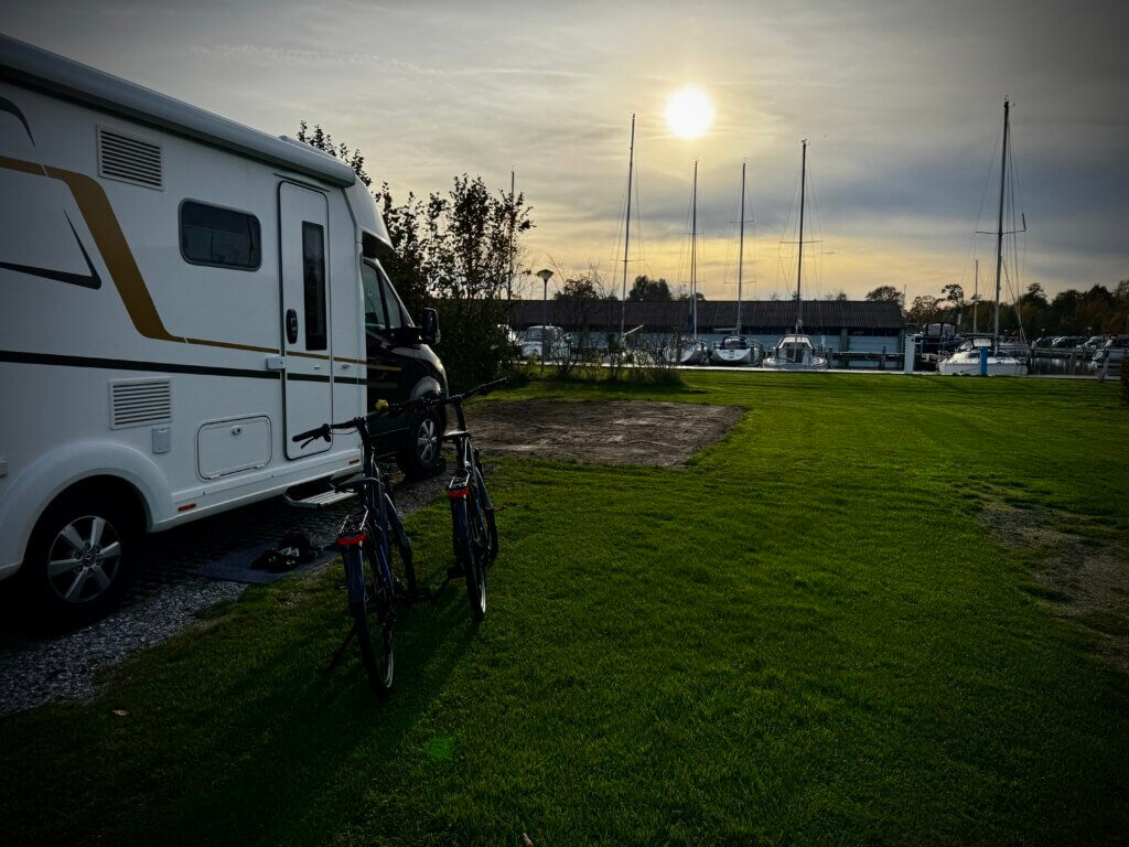 Wohnmobil auf einem grünen Stellplatz an der Marina in Sneek, mit zwei Fahrrädern davor und der Sonne tief am Himmel. Im Hintergrund liegen Segelboote am Steg. Ruhige, spätsommerliche Abendstimmung. Wohnmobilreise Sneek Niederlande.