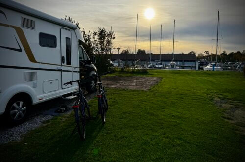Wohnmobil auf einem grünen Stellplatz an der Marina in Sneek, mit zwei Fahrrädern davor und der Sonne tief am Himmel. Im Hintergrund liegen Segelboote am Steg. Ruhige, spätsommerliche Abendstimmung. Wohnmobilreise Sneek Niederlande.